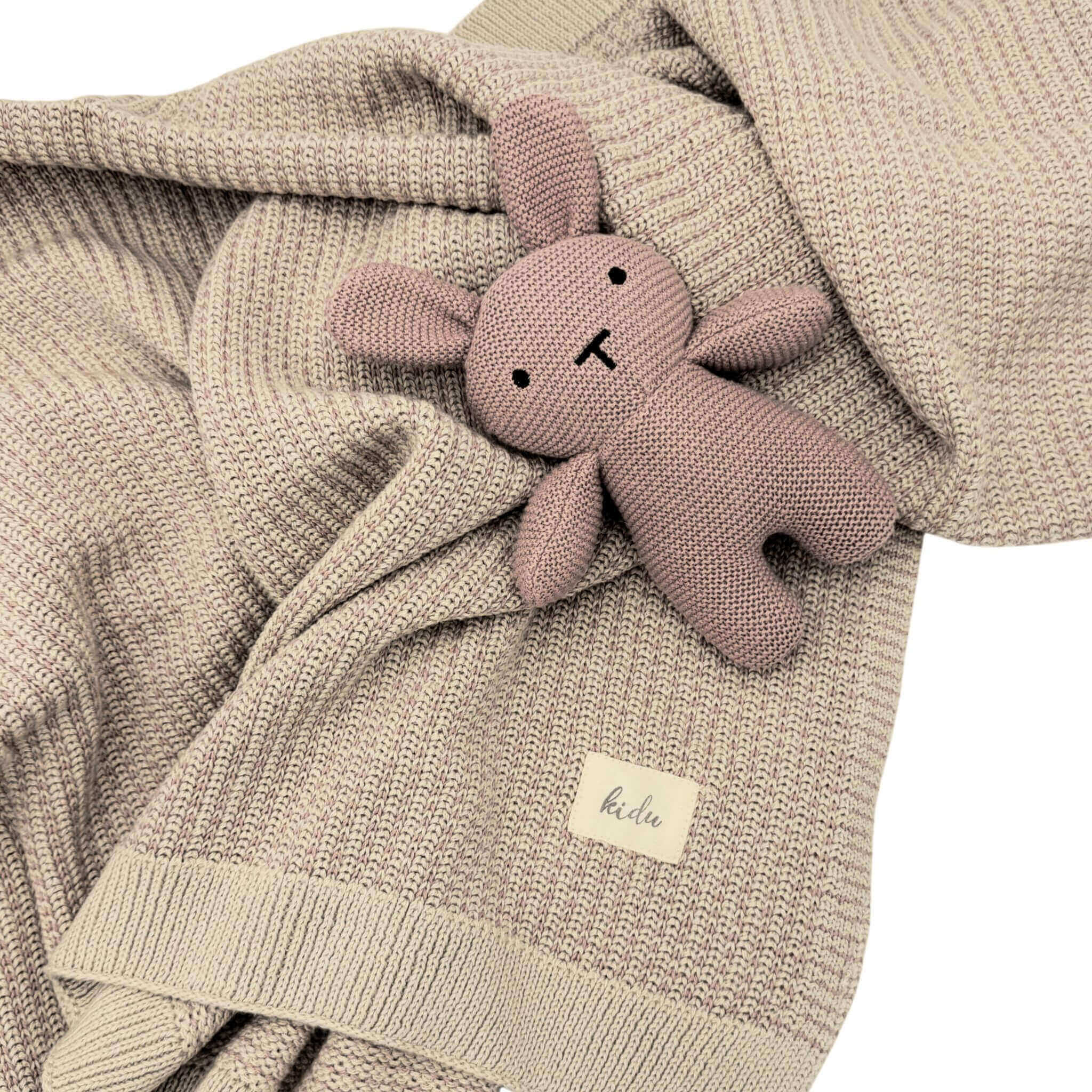 Pink knitted baby bunny toy on a beige and pink textured blanket with a visible brand label.